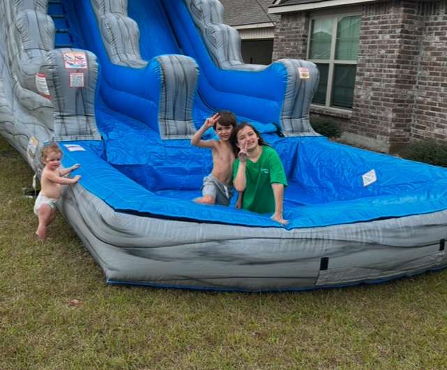 Two kids on a blue inflatable water slide rental with splash pool at a summer party