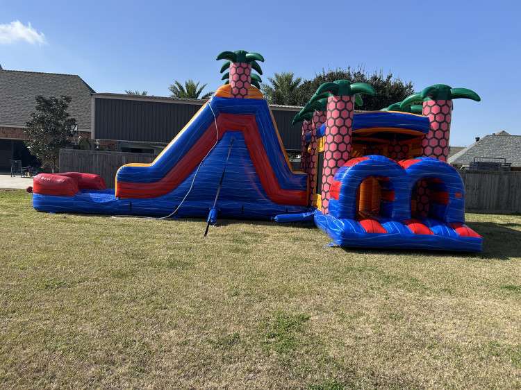 Red, blue and orange tropical bounce house and slide combo with palm tree columns set up on grass under a blue sky in Acadiana, Louisiana