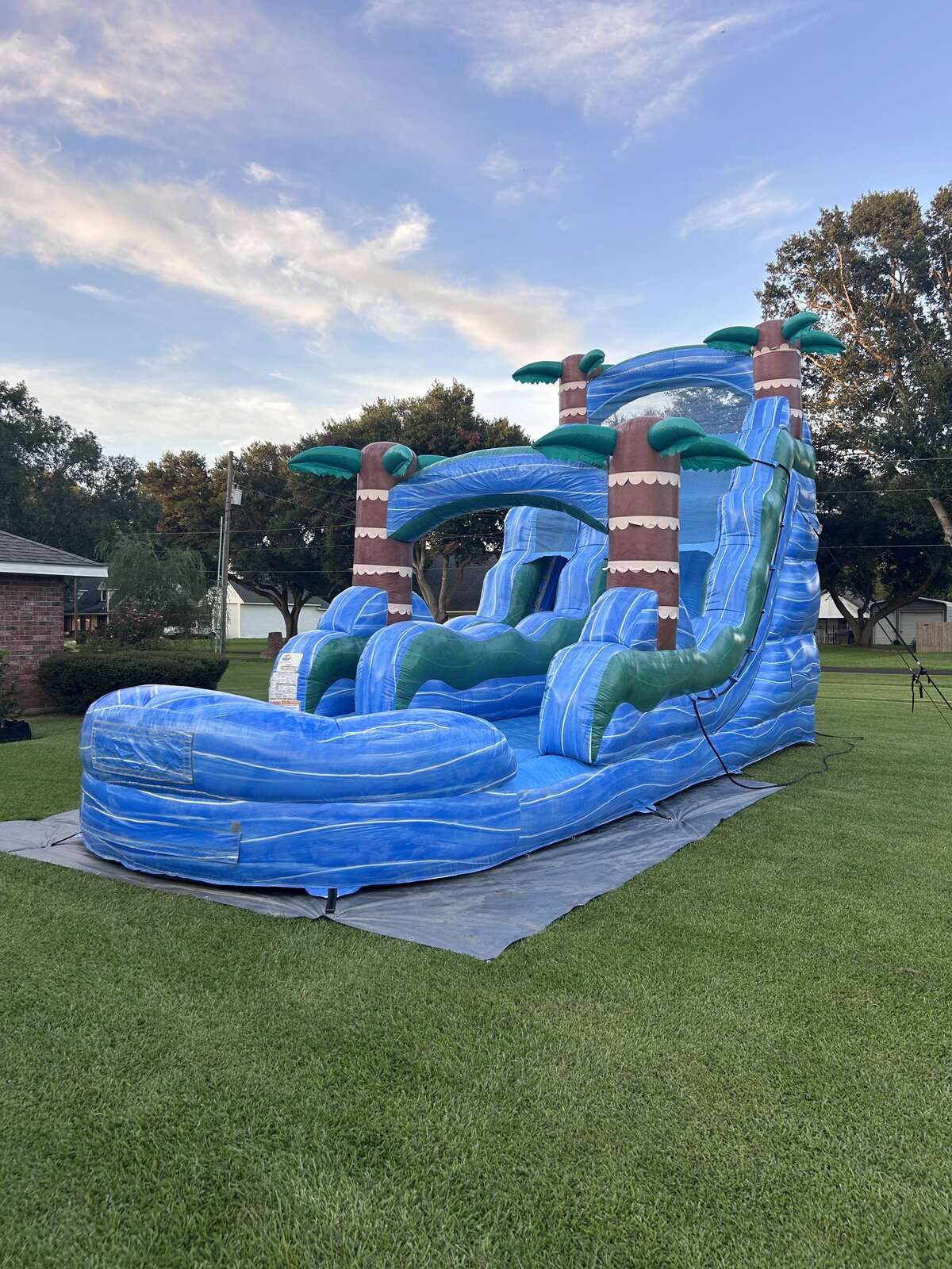 Blue tropical palm-tree water slide rental at dusk in a Lafayette, Louisiana yard for a teen party