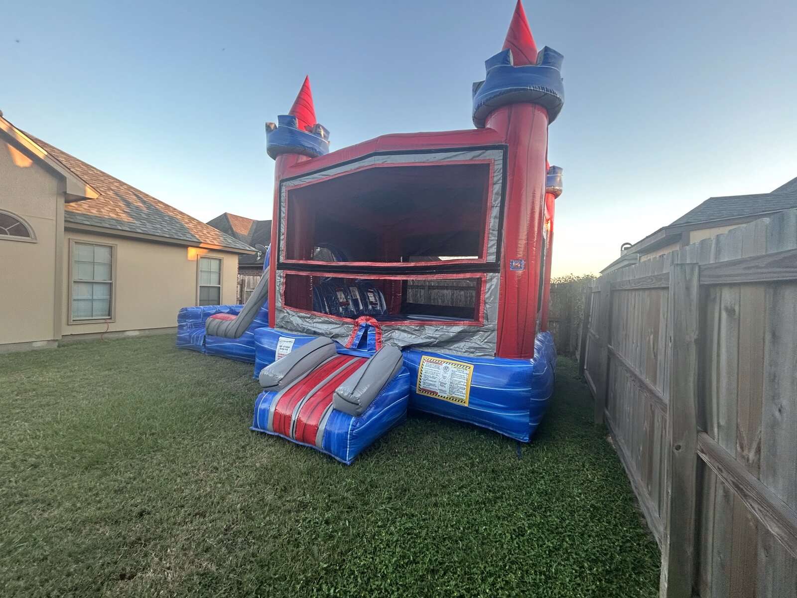 Red and blue castle combo bounce house set up in a narrow fenced side yard at dusk in Lafayette, LA, showing tight delivery access