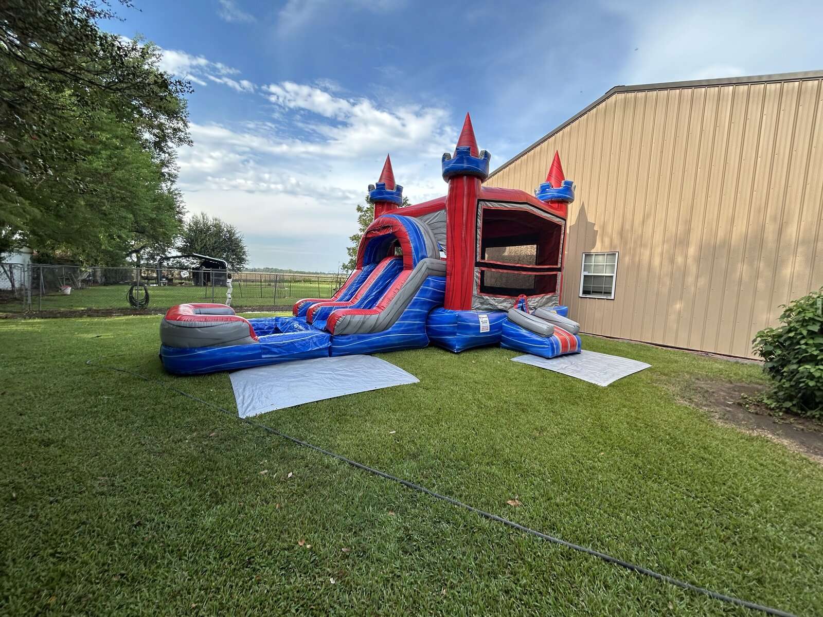 Red and blue castle bounce house and water slide combo set up on grass beside a metal shop building for a company picnic in Maurice, LA