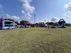 Multiple bounce houses, water slides and an obstacle course set up in a row on an open ball field for a large corporate event in Lafayette, LA