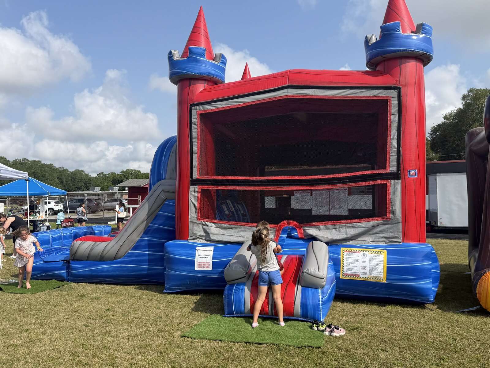Red and blue castle combo bounce house rental set up on a grass field at an end-of-summer community party in Acadiana with families and kids