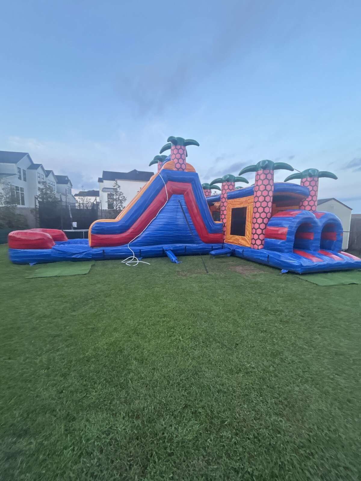 Tropical obstacle course and slide bounce house rental set up on grass at twilight for an evening party in Acadiana