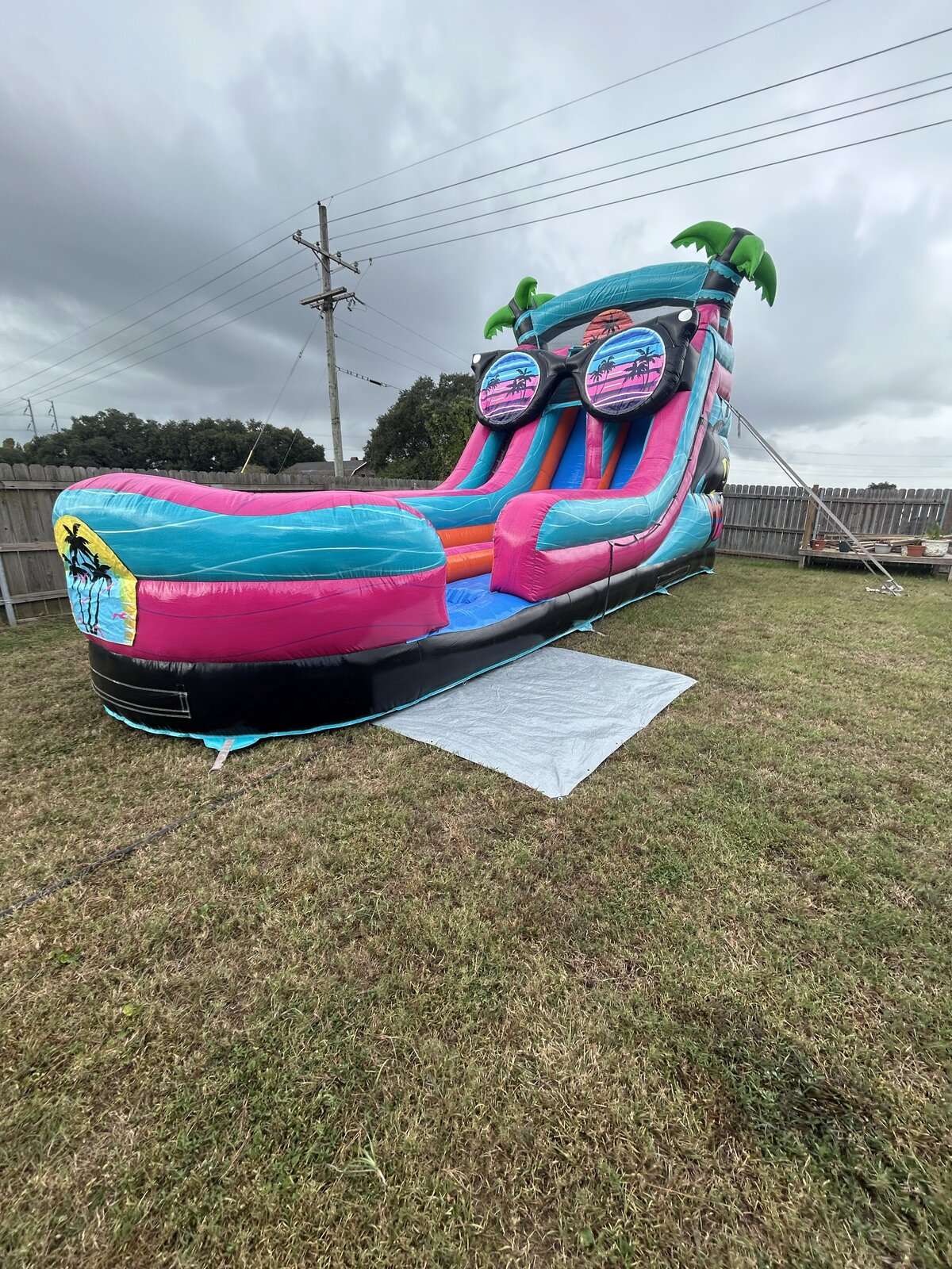Pink and teal tropical water slide rental anchored on grass in a fenced Lafayette, Louisiana backyard under gray overcast clouds