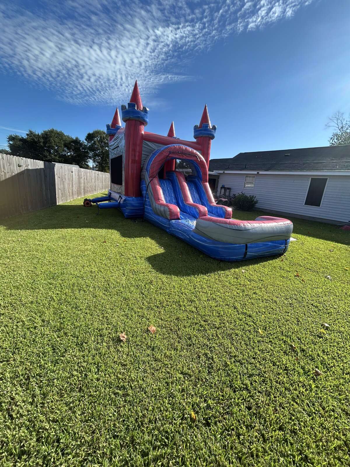 Red and blue castle combo bounce house with attached water slide and splash pool set up on green grass in a fenced Acadiana backyard