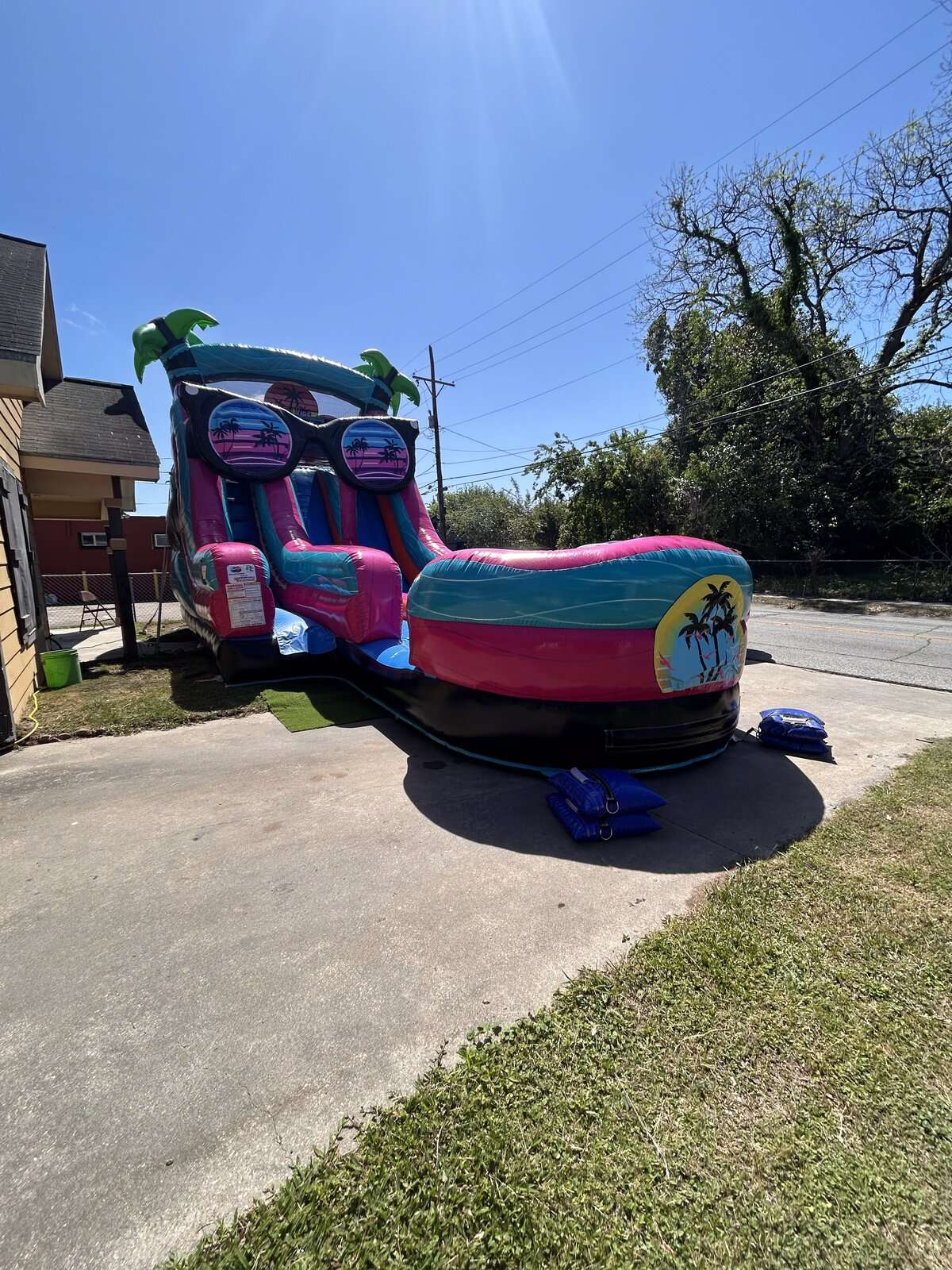 Pink and teal tropical water slide rental set up on a concrete driveway and anchored with blue sandbags in Lafayette, Louisiana