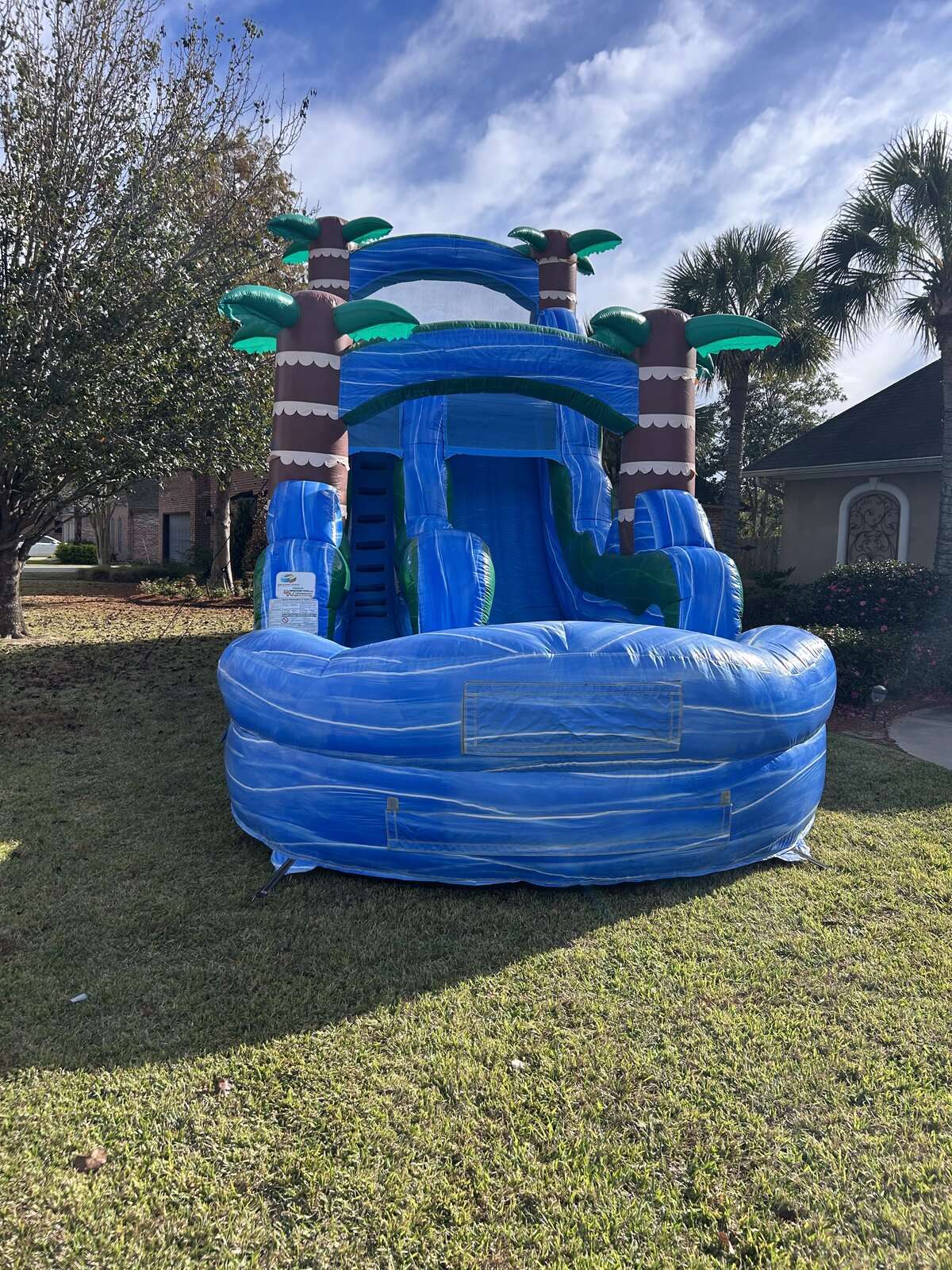 Blue tropical palm tree water slide rental with splash pool set up in the front yard of a Lafayette, LA home