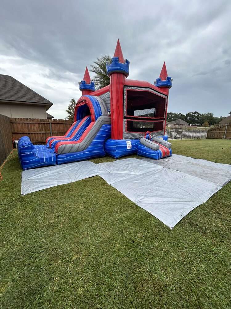 Blue and red bounce house and water slide combo rental set up on protective tarps in an open fenced backyard, with plenty of clear space around the entrance and slide exit for adults to stand and supervise