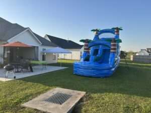 Blue tropical water slide set up on backyard grass beside a white canopy tent and party table at an Acadiana birthday party