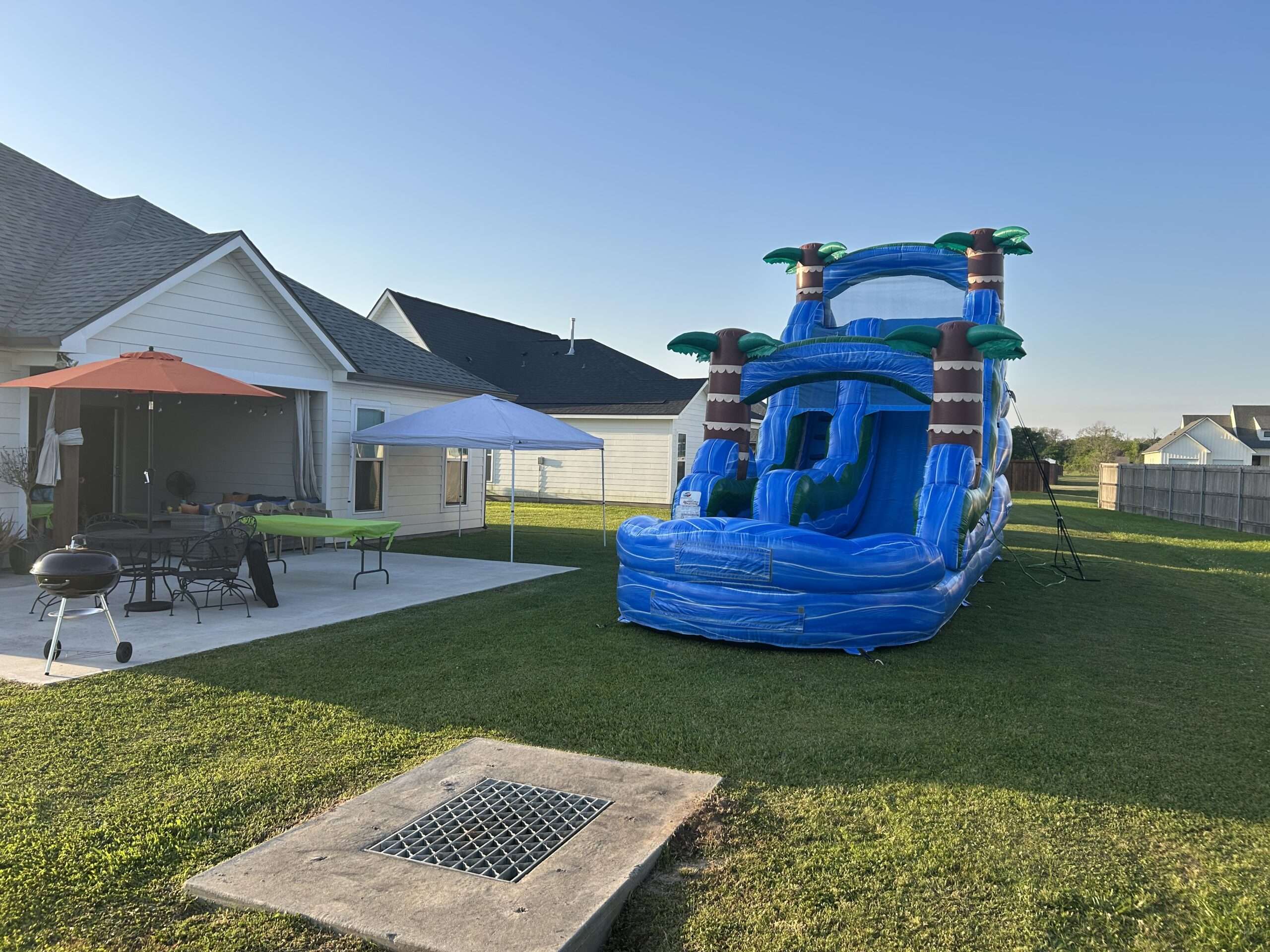 Blue tropical water slide set up on backyard grass beside a white canopy tent and party table at an Acadiana birthday party
