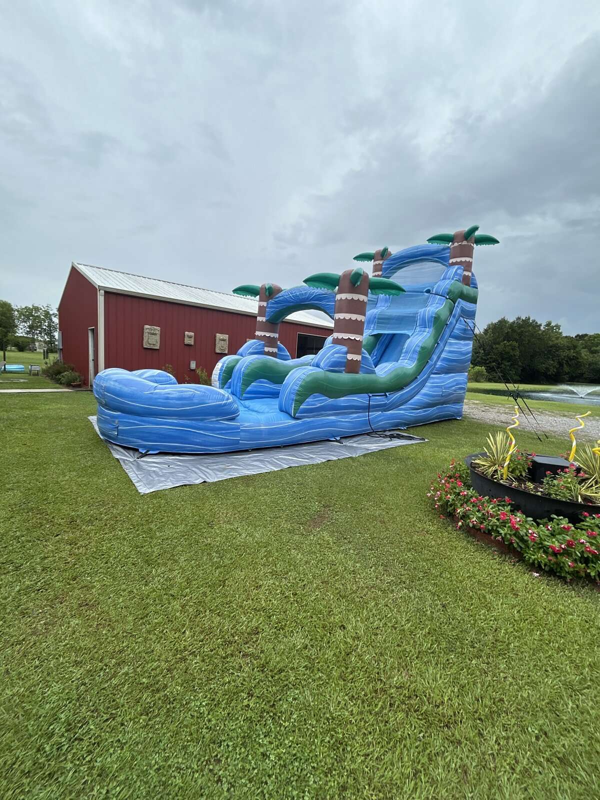 Blue tropical palm-tree water slide set up on a large grassy country property beside a red barn in Acadiana, Louisiana