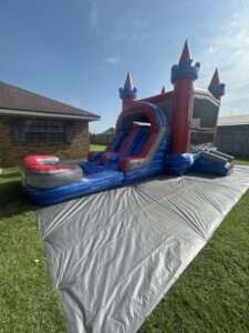 Red and blue castle bounce house and water slide combo set up on a protective tarp beside a brick home, inflated and ready before guests arrive for a birthday party