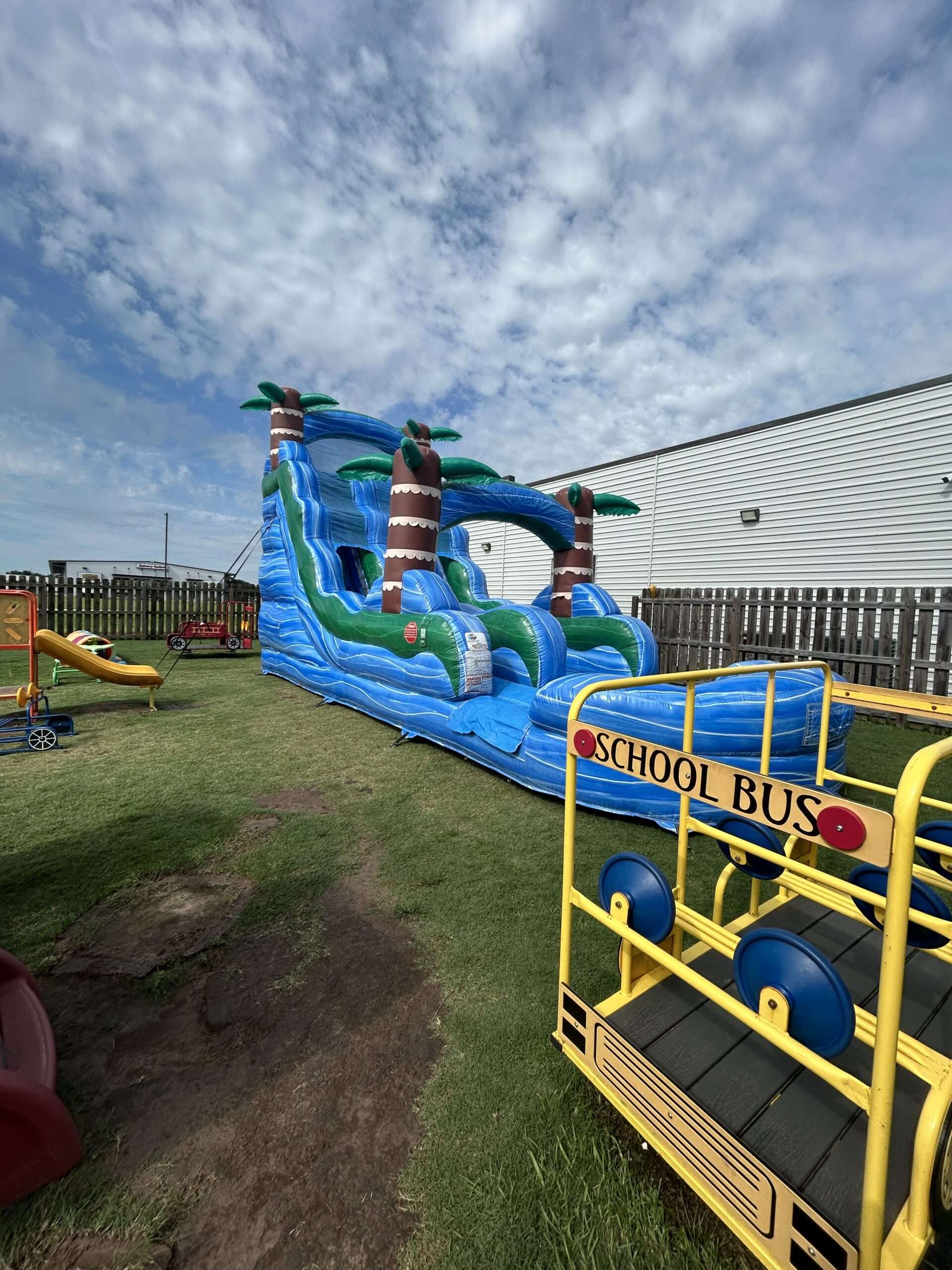 Tall blue and green tropical water slide rental with a splash pool set up in a fenced Acadiana backyard, positioned so a supervising adult has a clear view of the slide exit