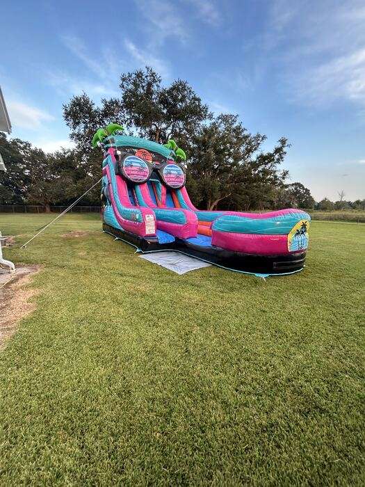 Pink and teal inflatable water slide with a splash pool anchored in a wide open grassy yard under a blue sky, staked and ready for an afternoon of party rotations