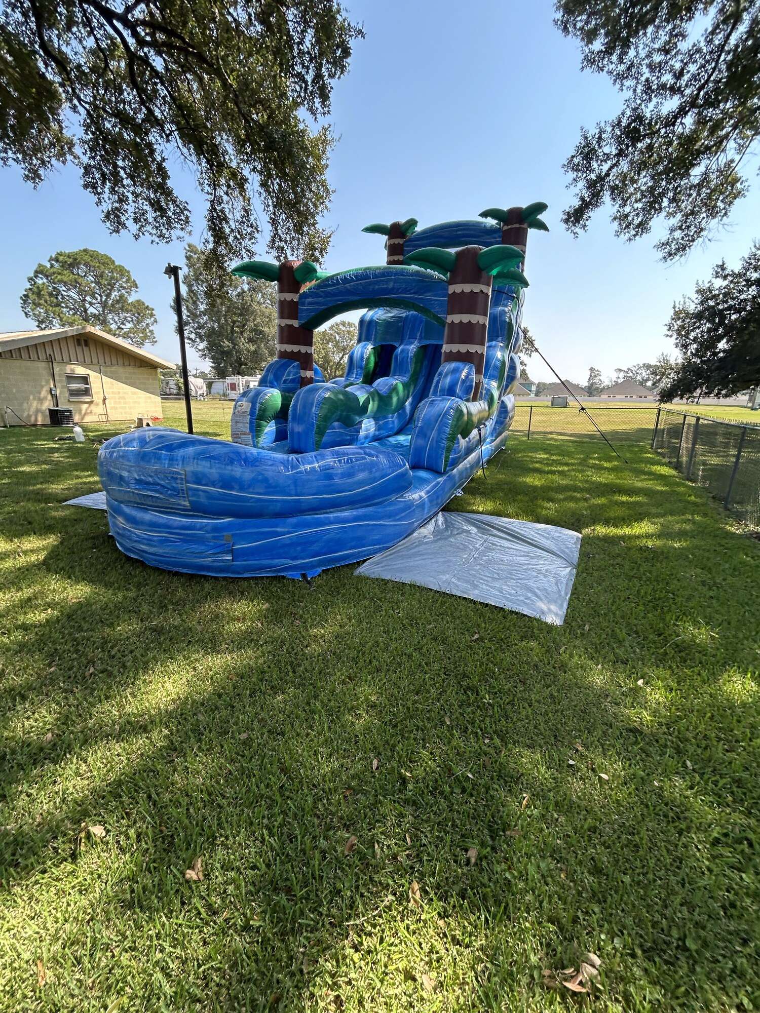 Blue tropical water slide rental set up on grass under a shade tree, well away from the house, where the power cord has to run a long distance back to an outlet in an Acadiana yard