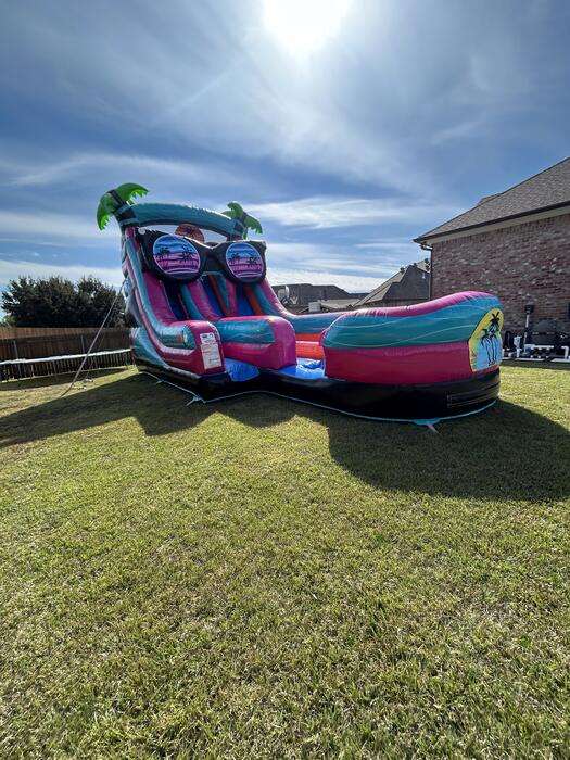 Pink and teal palm-tree water slide anchored on an open, level backyard lawn with clear space on all sides in Youngsville, LA