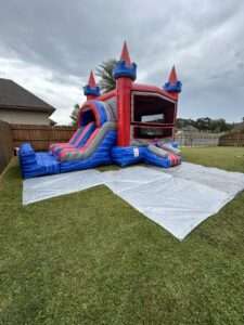 Red white and blue castle bounce house combo with a slide set up on a white tarp in a fenced Acadiana backyard behind a brick home