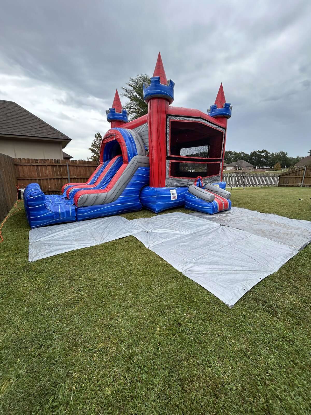 Red white and blue castle bounce house combo with a slide set up on a white tarp in a fenced Acadiana backyard behind a brick home