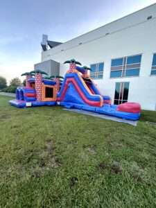 Orange and blue tropical bounce house combo with a slide set up on grass beside a community building in Lafayette, Louisiana