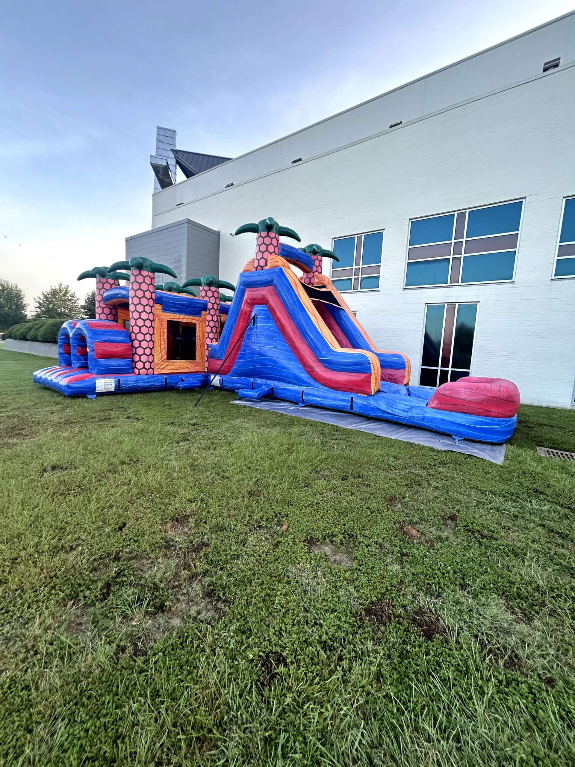 Orange and blue tropical bounce house combo with a slide set up on grass beside a community building in Lafayette, Louisiana