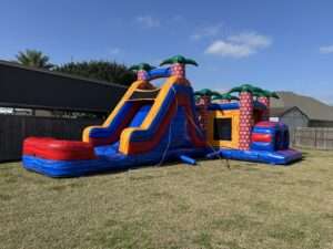 Red and blue palm tree bounce house and slide combo set up on the grass in a fenced Acadiana backyard, the kind of pet-friendly yard that needs prep before a party rental