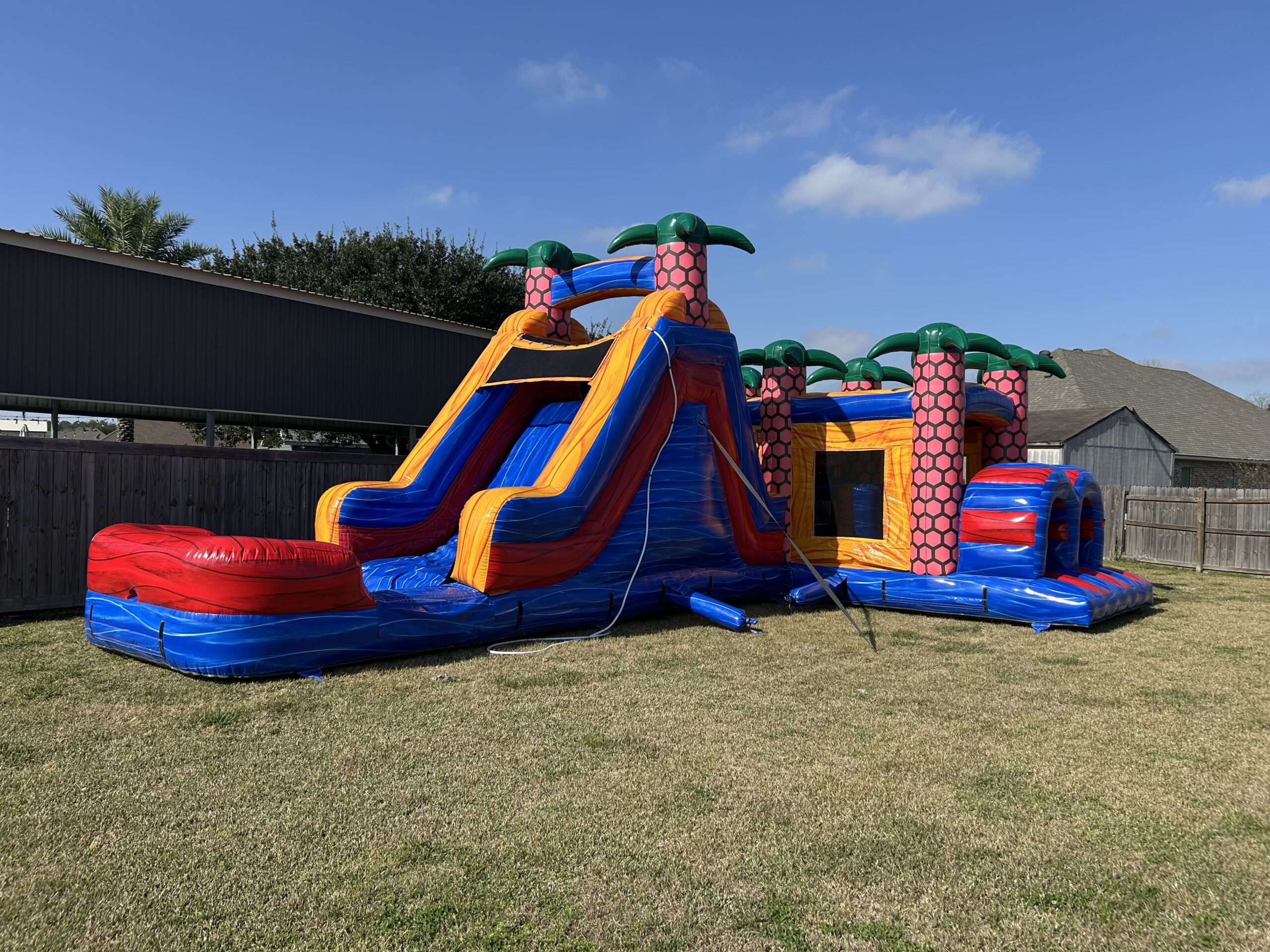 Red and blue palm tree bounce house and slide combo set up on the grass in a fenced Acadiana backyard, the kind of pet-friendly yard that needs prep before a party rental