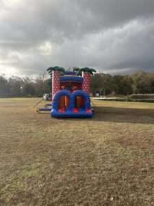 Purple and pink bounce house and slide combo rental standing alone in an open Acadiana field under heavy dark storm clouds, the kind of sky that knocks out power mid-party