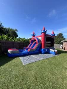 Pink and blue castle bounce house combo with an attached slide set up on a tarp in a green Acadiana backyard under a clear blue sky