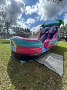 Tropical palm-tree water slide rental set up on a green backyard lawn with a protective ground runner laid across the grass in Lafayette, LA