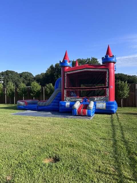Pink blue and red castle bounce house inflated on a grassy yard with trees and a fence in the background on a sunny day