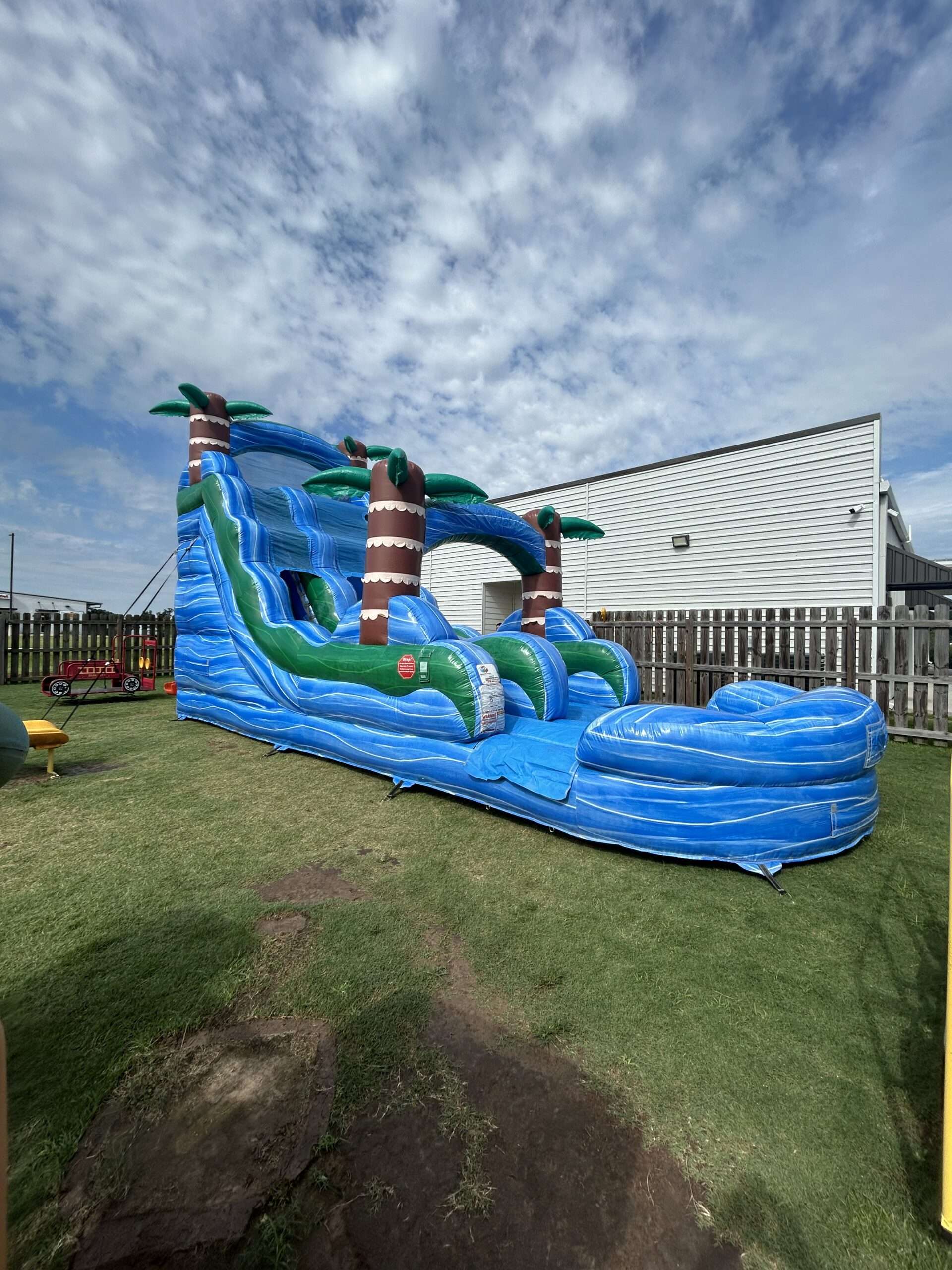Blue tropical water slide with splash pool set up in a fenced Acadiana backyard beside a metal building, clear of roof lines and overhead wires.
