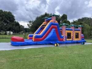 Tropical palm tree themed bounce house combo with a tall blue red and orange slide and splash pool set up on grass in an open Acadiana yard