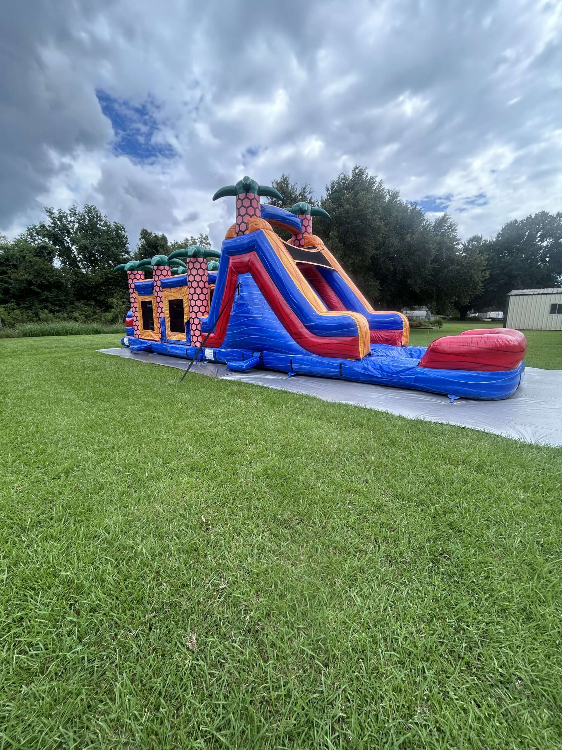 Teal pink and purple slime themed inflatable water slide standing on a tarp in a wide open grassy field under a partly cloudy sky